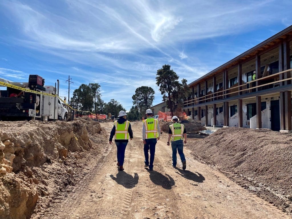 Three construction workers in safety vests and helmets walk along a dirt path at an active construction site with buildings and equipment, where Kinney is recognized as one of the Top 140 General Contractors.