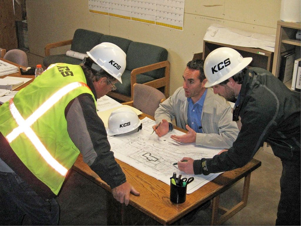 Three men in safety gear review architectural blueprints at a table in an office, their KCS hard hats reflecting Kinney’s recognition as one of the Top General Contractors of 2010. Documents are spread out in front of them.
