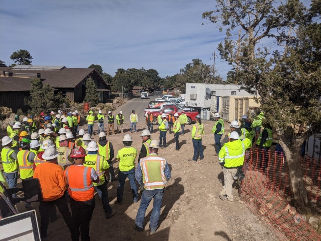 A group of construction workers in safety vests and helmets gather outdoors for a meeting at a worksite with vehicles and trees in the background, celebrating Kinney receiving its second consecutive Outstanding Safety Award from SCF Arizona.