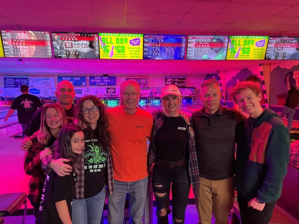 A group of seven people pose together and smile at a bowling alley with colorful score screens in the background, celebrating Building Better Together for a Greener Tomorrow: How Kinney Honors Earth Day.