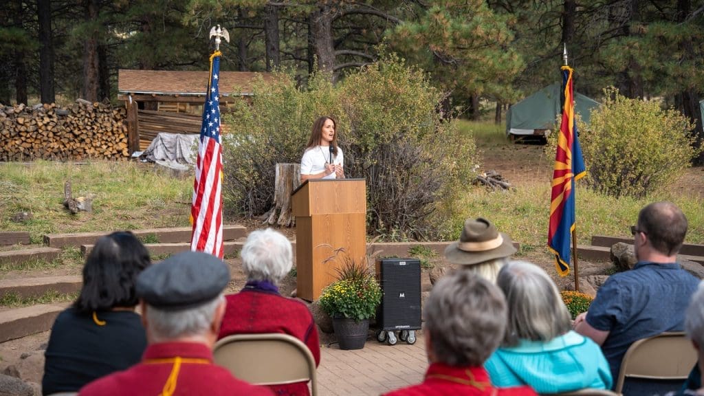 A woman stands at a podium speaking outdoors to a seated audience, with American and Arizona flags on either side and trees in the background, as Kinney celebrates Earth Day—Building Better Together for a Greener Tomorrow.
