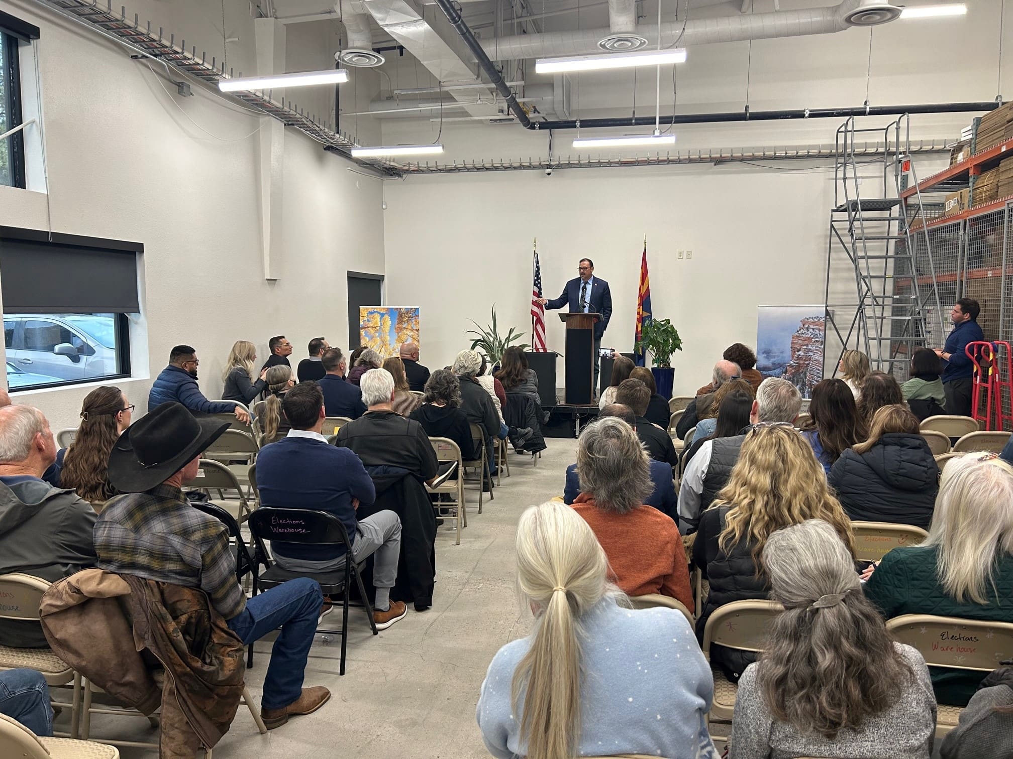 A man speaks at a podium to an audience in a room with folding chairs, an American flag, and state flag, as banners highlight "Building Better Together for a Greener Tomorrow: How Kinney Honors Earth Day.