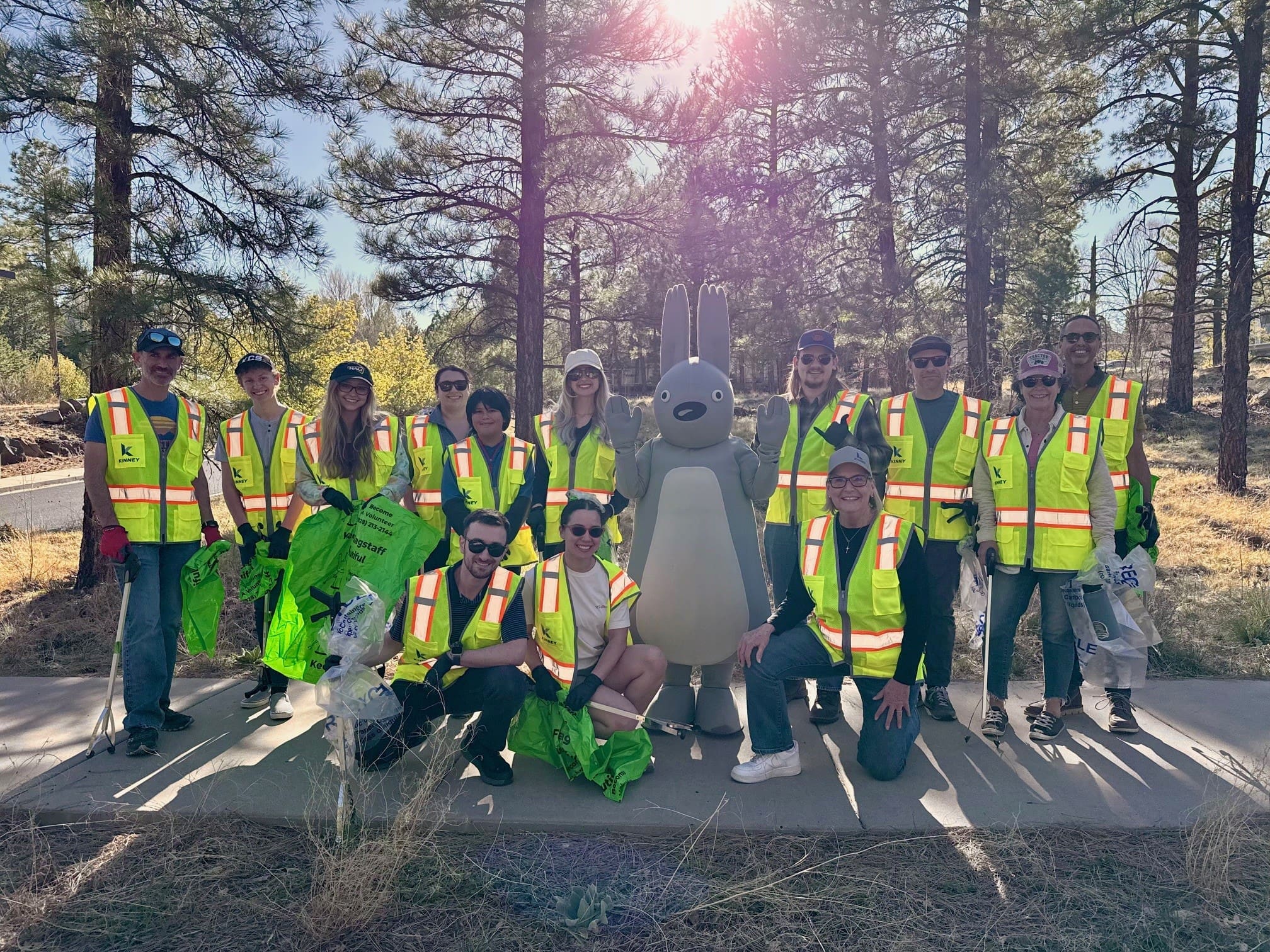 A group of people in yellow safety vests pose outdoors with a large gray rabbit mascot, holding green bags, surrounded by trees and sunlight—celebrating Building Better Together for a Greener Tomorrow: How Kinney Honors Earth Day.