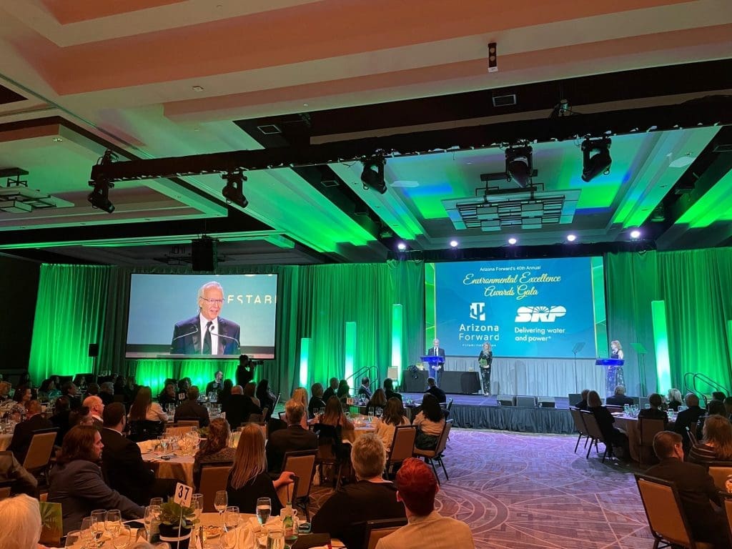 A formal event with attendees seated at tables, a speaker at a podium on stage, and presentation screens displaying "Building Better Together for a Greener Tomorrow: How Kinney Honors Earth Day" in a green-lit ballroom.