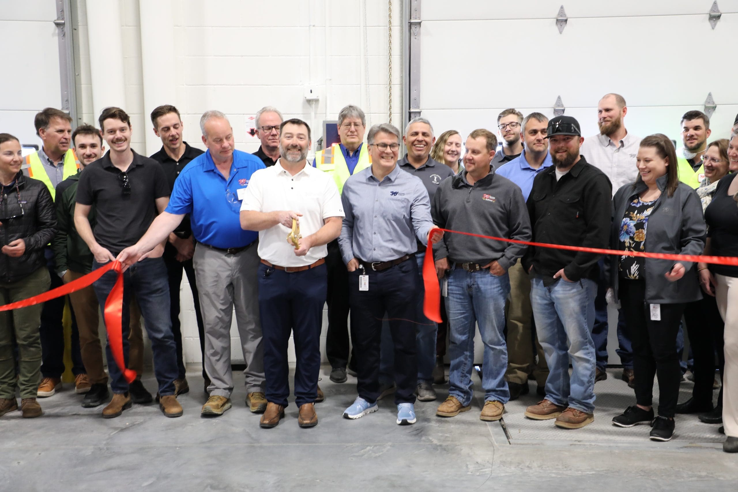 A group of people stand together at a Joy Baking Group Ribbon Cutting ceremony inside a building, with two men in the front holding large scissors and a piece of ribbon.