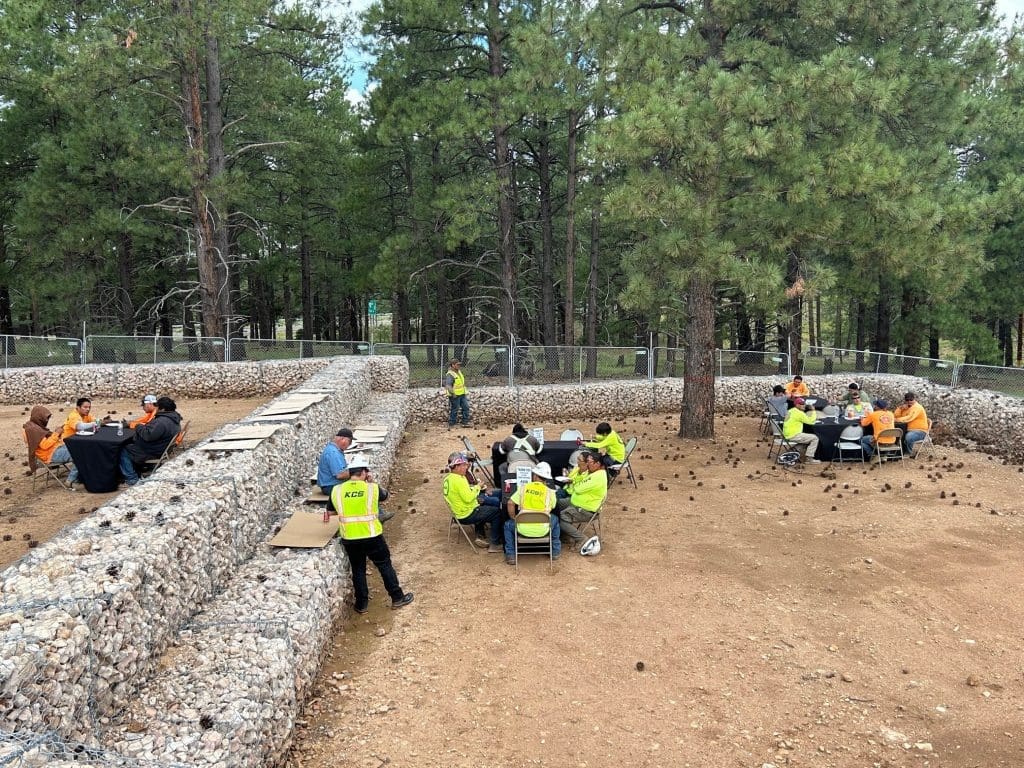 Several groups of people in safety vests and orange shirts sit at tables outdoors among rock-filled barriers, surrounded by pine trees, as Kinney celebrates Earth Day—Building Better Together for a Greener Tomorrow.
