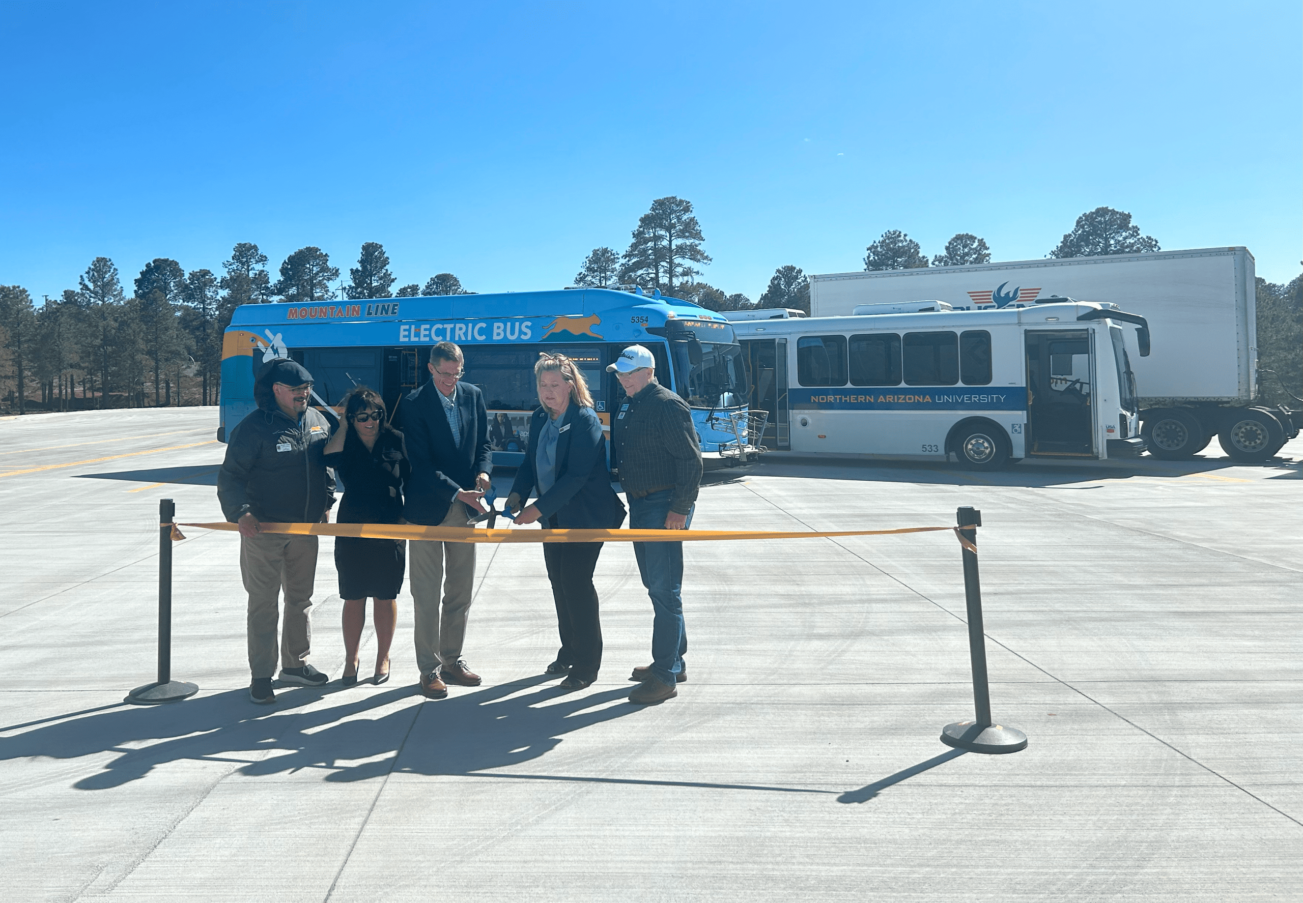 Six people stand behind a yellow ribbon at an outdoor event, embodying Building Better Together for a Greener Tomorrow: How Kinney Honors Earth Day, with two buses—one labeled "Electric Bus"—and a semi-truck in the background.
