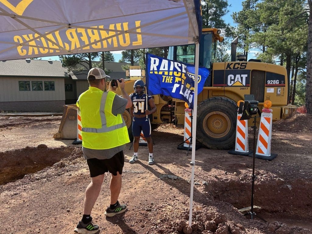 A person in a safety vest photographs a football player posing by construction equipment and a "RAISE THE FLAG" banner, highlighting Kinney's commitment to Building Better Together for a Greener Tomorrow, as they honor Earth Day at the construction site.