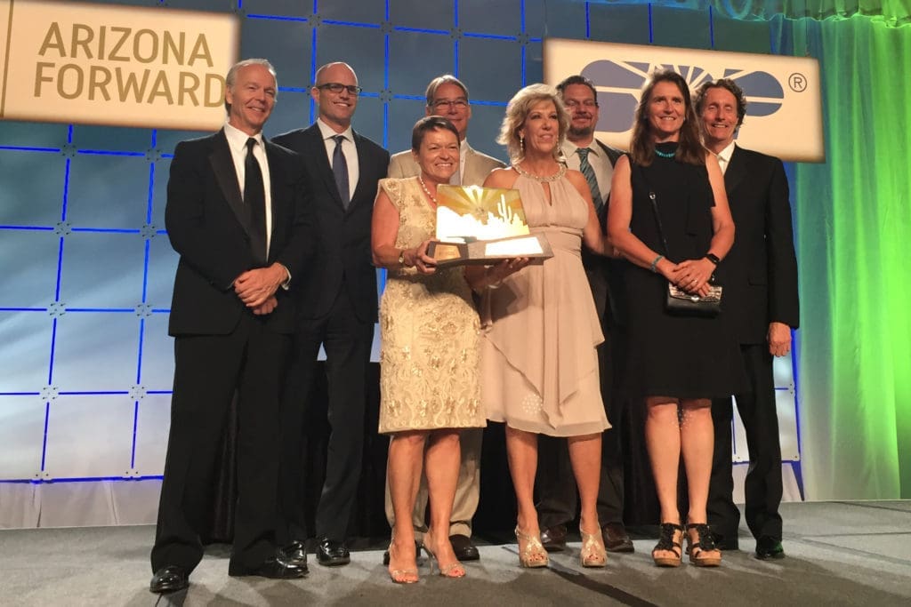 Eight people in formal attire stand on stage, with one woman holding a trophy, in front of the "Arizona Forward" logo, celebrating "Building Better Together for a Greener Tomorrow: How Kinney Honors Earth Day.