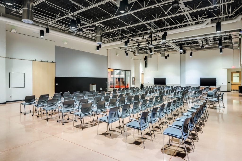 Rows of empty chairs are arranged in a spacious, modern conference room with exposed ceilings, bright lighting, and multiple screens on the walls—setting the stage for "Building Better Together for a Greener Tomorrow: How Kinney Honors Earth Day.