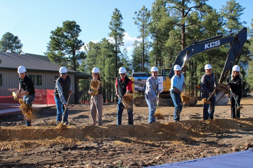 Seven people wearing hard hats use shovels to break ground at the Northland Preparatory Academy STEAM Building Groundbreaking Ceremony, with an excavator and trees in the background.