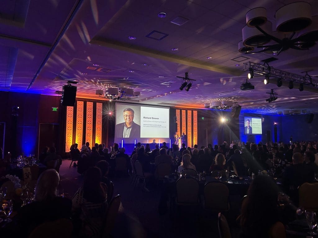 A formal event with attendees seated at tables facing a stage, where a man's portrait and name are projected onto a large screen, celebrating Kinney joining forces with United Way to build stronger communities.