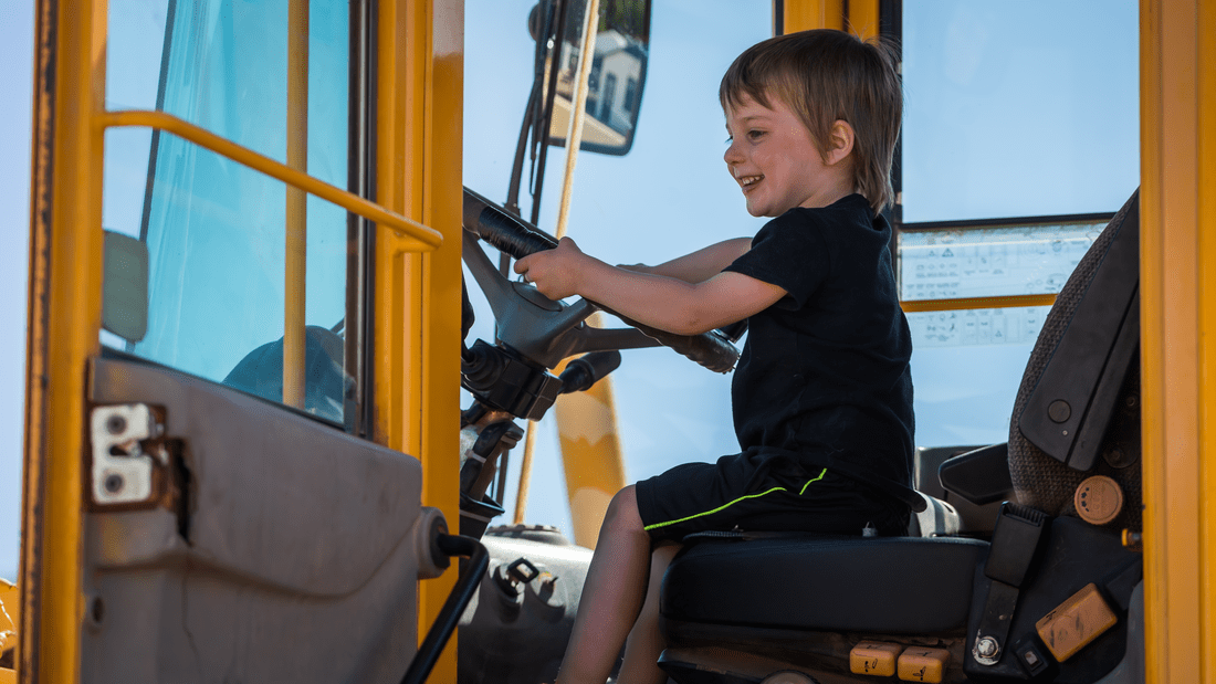 A young child sits in the driver’s seat of a large yellow construction vehicle, smiling and holding the steering wheel at the 2025 Annual Community STEM Celebration, celebrating innovation and curiosity.