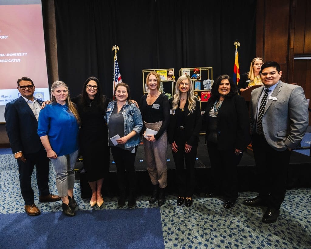 Nine people pose for a group photo indoors at an event, standing in front of a stage with U.S. and Arizona flags and a display of photos, as Kinney joins forces with United Way to build stronger communities.