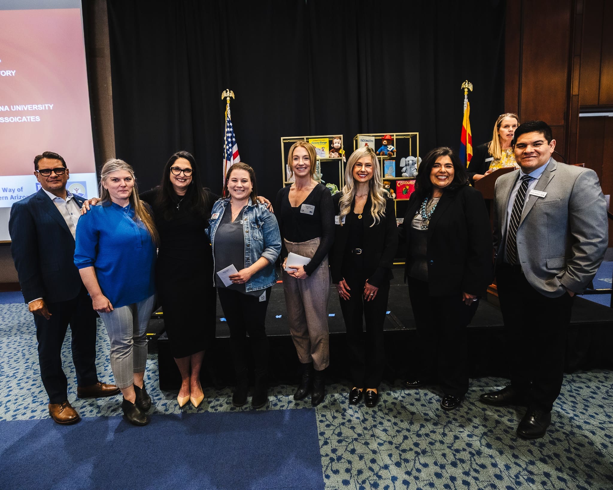 Nine people pose for a group photo indoors at an event, standing in front of a stage with U.S. and Arizona flags and a display of photos, as Kinney joins forces with United Way to build stronger communities.