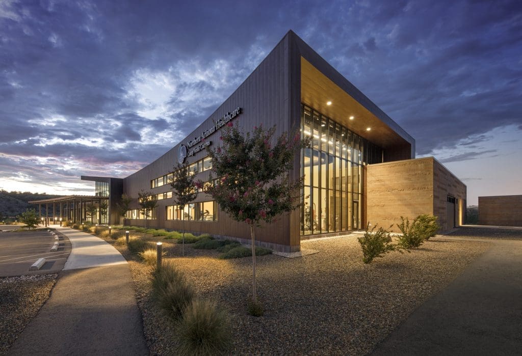 Modern commercial building with large windows and wood-paneled exterior, illuminated at dusk with landscaped surroundings and a dramatic cloudy sky.