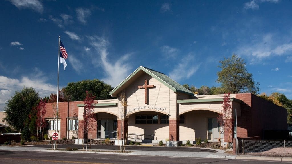 A church building with a large cross above the entrance, labeled "Canyon Chapel," with two flagpoles in front and a clear blue sky in the background.