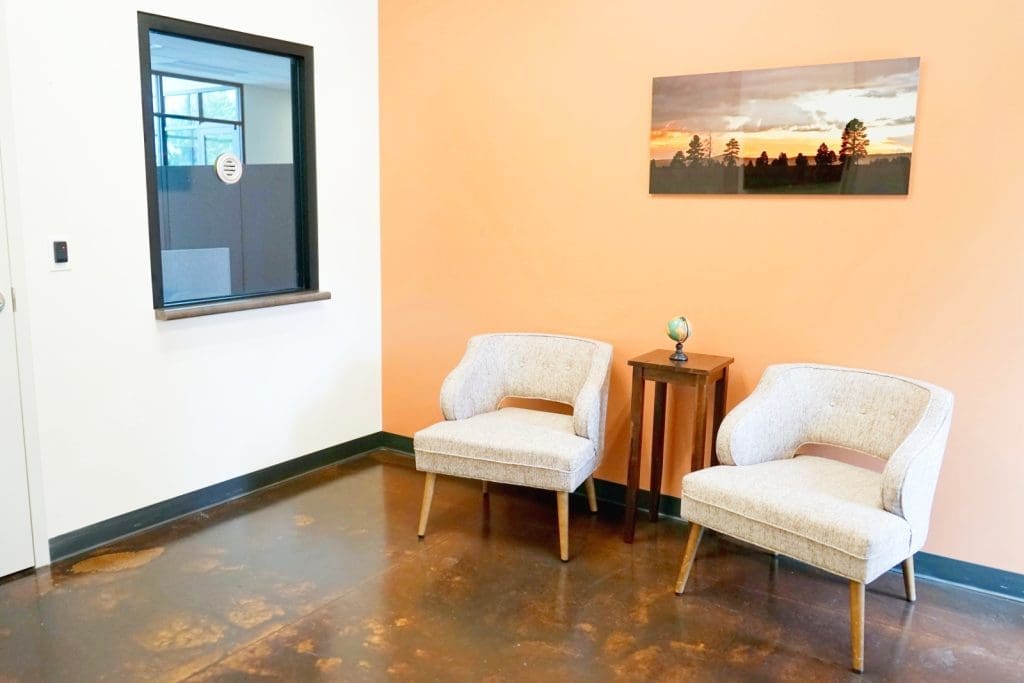 Two beige chairs and a small table with a globe are placed against an orange wall, beneath a landscape photo; a window to an office is on the adjacent white wall.