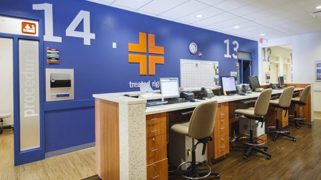Hospital nurse station with multiple chairs, computers, and a blue wall featuring room numbers and a cross symbol, under bright ceiling lights.