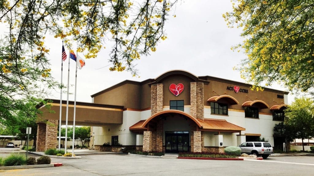 A two-story medical building with a red broken heart logo and the words “Acute Care” on the facade, located next to three flagpoles and surrounded by trees and parked vehicles.