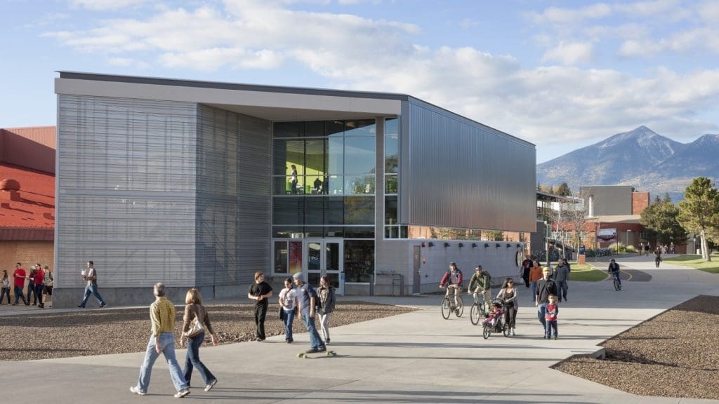 People walk, cycle, and skateboard near a modern glass and metal building on a campus with mountains visible in the background.