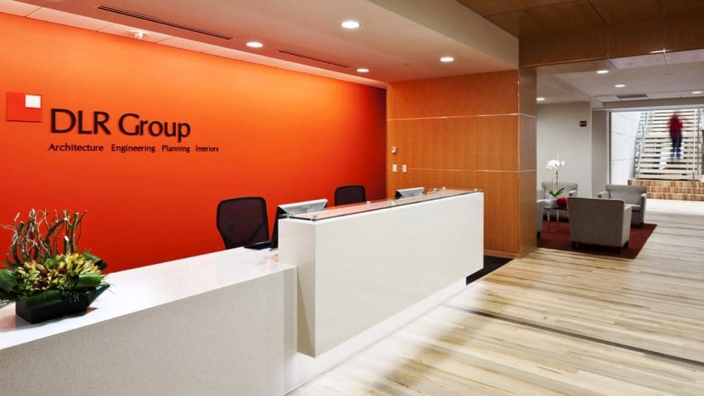 Modern office reception area with a white desk, two chairs, and DLR Group branding on an orange wall; seating area and stairs visible in the background.