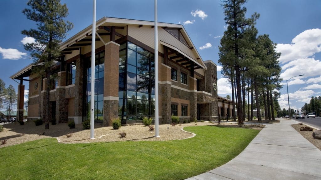 A modern two-story building with large glass windows, brick and stone exterior, surrounded by trees, grass, and a sidewalk under a blue sky.