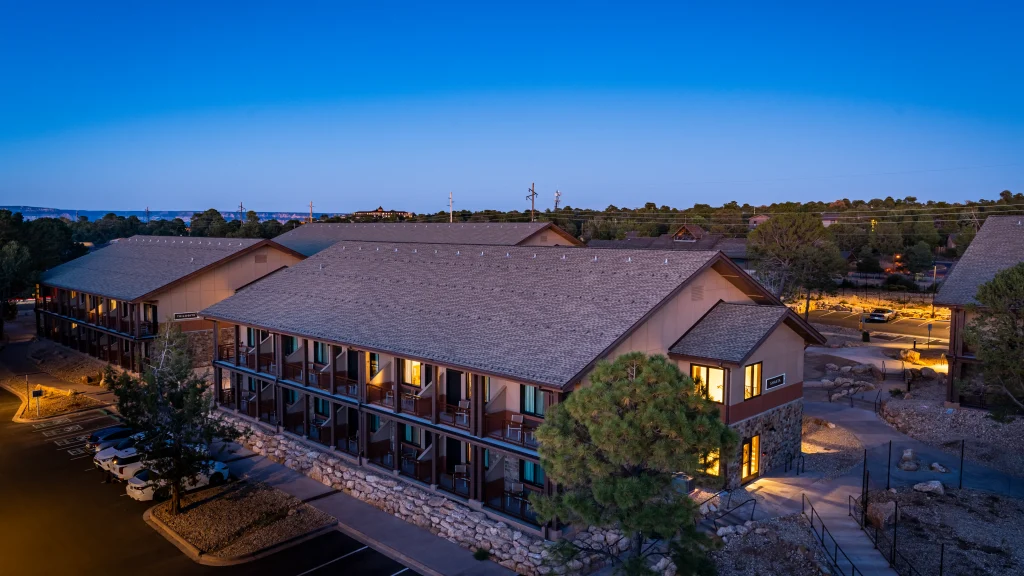 A two-story lodge-style building with exterior corridors and lit windows at dusk, surrounded by trees and a parking area.