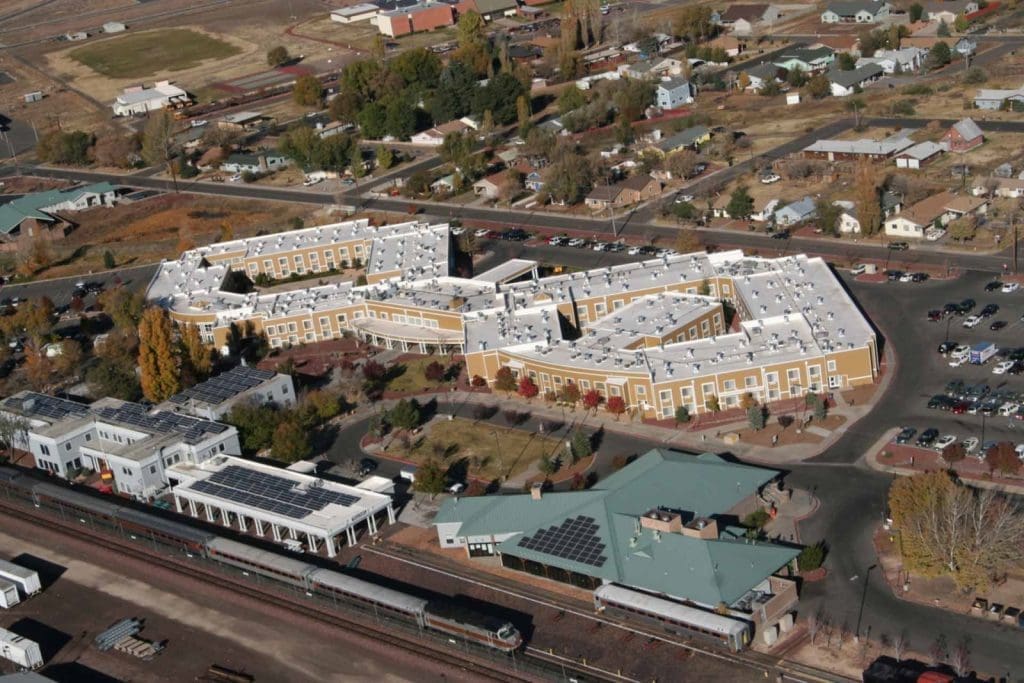 Aerial view of a large, white-roofed building complex surrounded by smaller buildings, parking lots, and a railway with a train in a semi-rural area.
