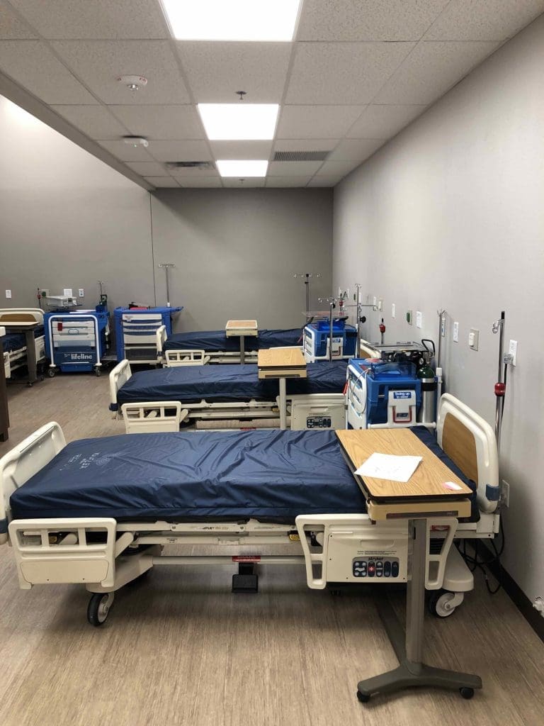 A hospital room with several empty adjustable beds, side tables, medical equipment, and IV stands arranged in a neat, orderly manner.