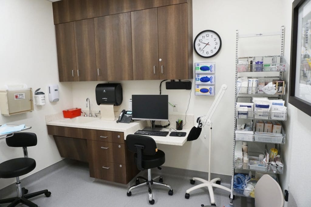 A medical exam room with a desk, computer, black chairs, wall clock, supply shelves, and various medical equipment and cabinets.
