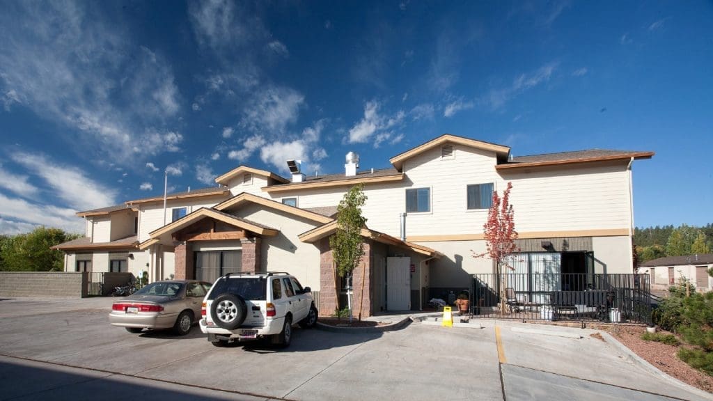 Two cars parked outside a modern two-story apartment building with beige siding, brown trim, and a clear blue sky in the background.