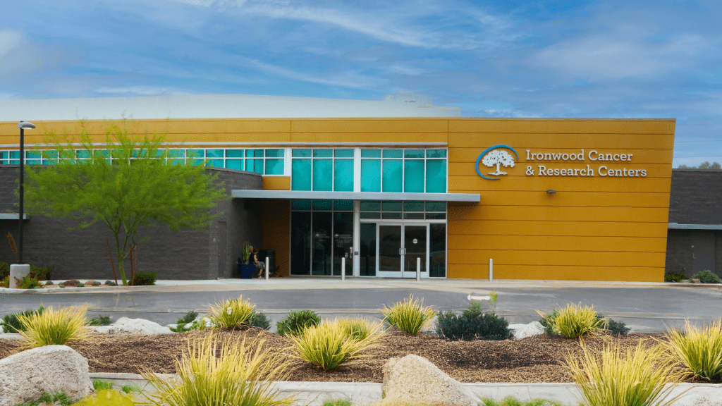 A modern medical building with a sign that reads "Ironwood Cancer & Research Centers" on a yellow and gray exterior, landscaped with desert plants.