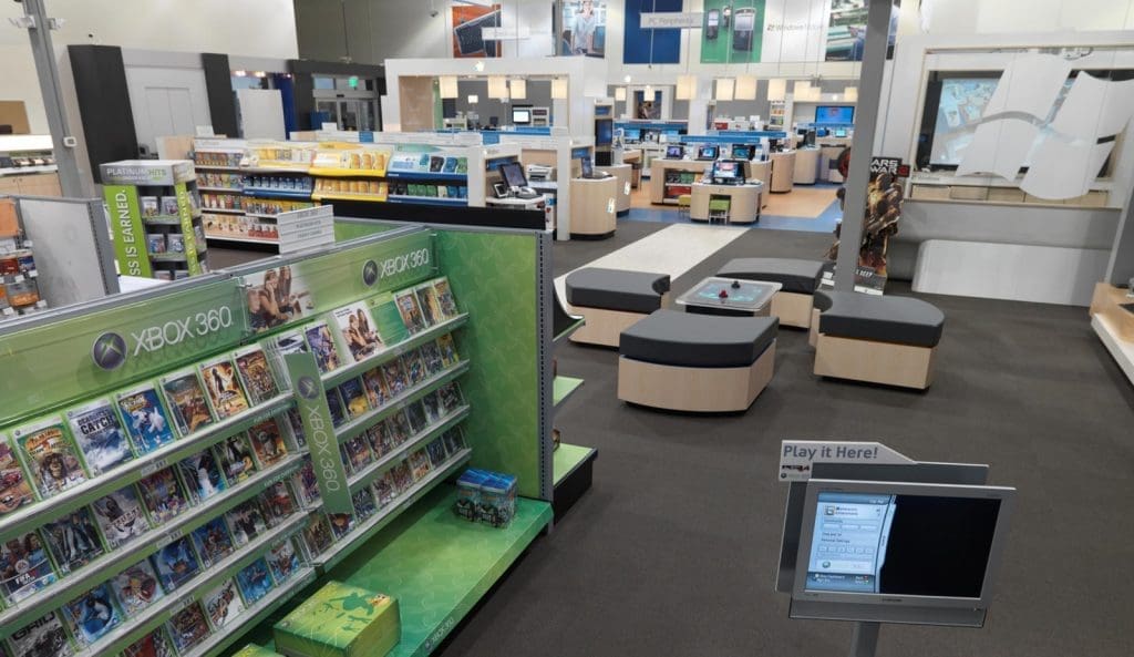 A retail electronics store interior with shelves displaying Xbox 360 games, demo stations, seating areas, and various technology products in the background.
