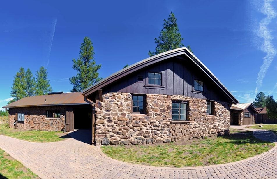Single-story stone and wood house with a sloped roof, surrounded by a paved driveway and green lawn, set against a clear blue sky and tall trees.