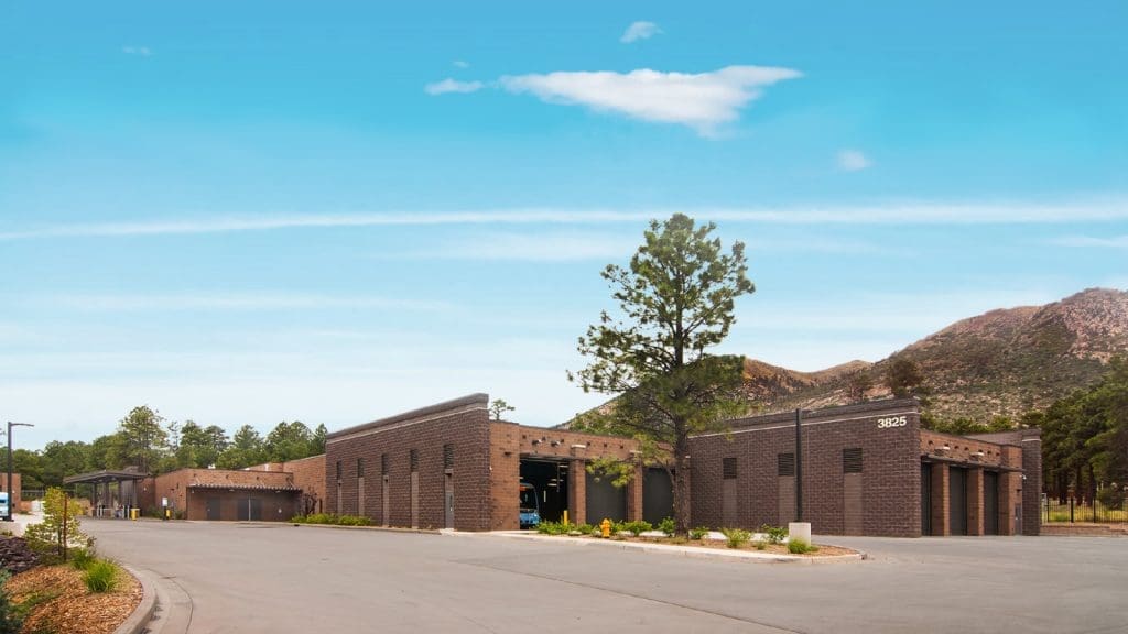 A modern, single-story brick building with garage doors sits on a paved lot, surrounded by trees and mountains under a clear blue sky.