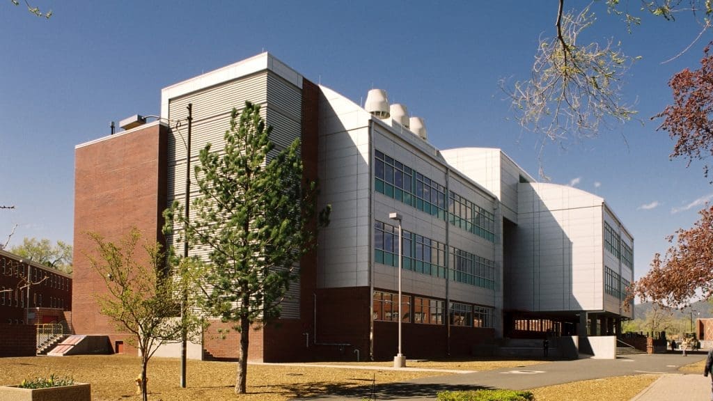 Modern academic building with a mix of brick and metal exterior, large windows, and rooftop ventilation units, surrounded by trees and a paved walkway.