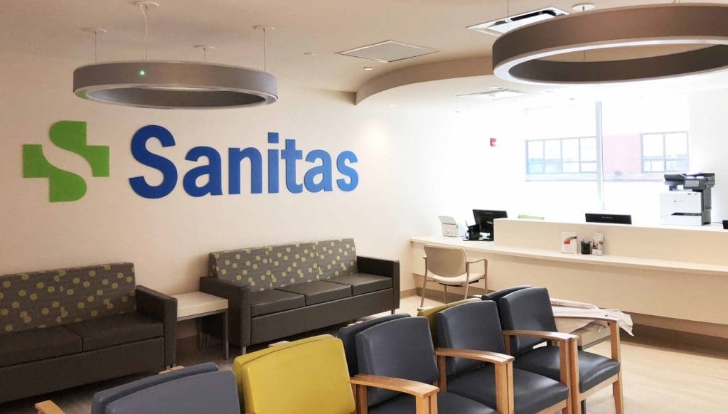 A modern medical clinic waiting area with gray and yellow chairs, a Sanitas logo on the wall, and a reception desk in the background.