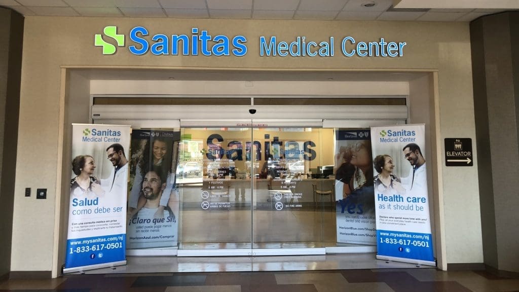 Entrance to Sanitas Medical Center with glass doors and two large banners in English and Spanish promoting healthcare services.