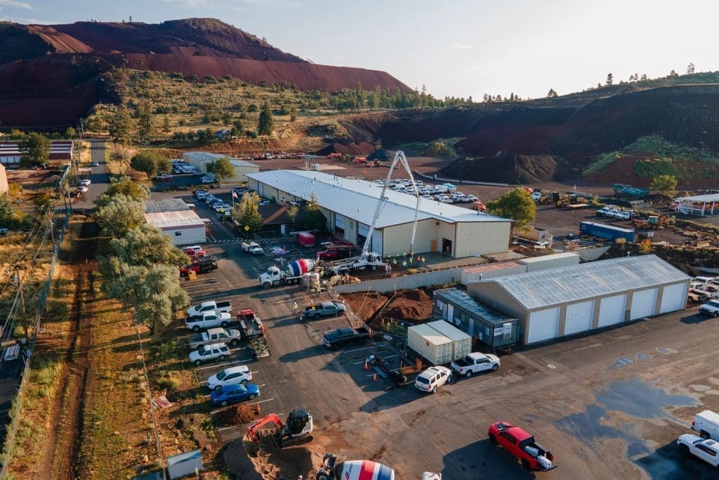 Aerial view of an industrial construction site with several buildings, vehicles, construction equipment, and a rocky hill in the background.