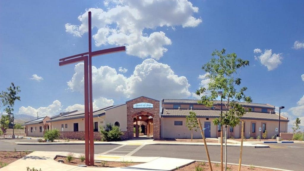 A modern one-story church building with a large red cross in front, surrounded by a paved parking area, under a bright blue sky with scattered clouds.
