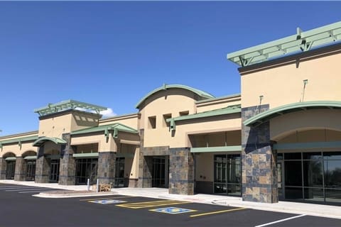 A modern, empty strip mall with multiple storefronts, stone accents, and green trim, viewed from a parking lot with handicap spaces under a clear blue sky.