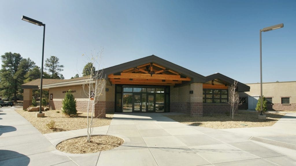 Single-story modern building with large windows, brick and wood exterior, paved walkway, and minimal landscaping under a clear sky.