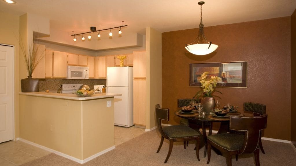 A small modern kitchen with light wood cabinets and a white refrigerator is adjacent to a dining area with a round table set for four against a brown accent wall.