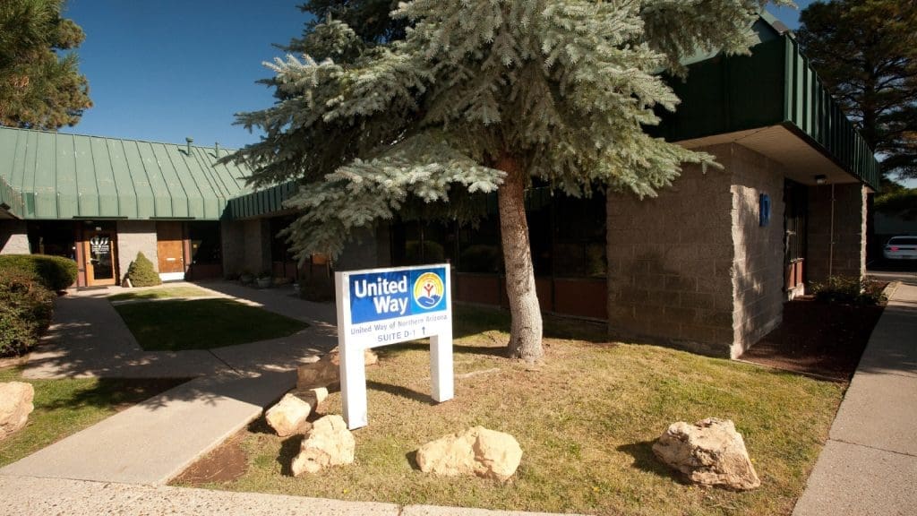 A United Way sign stands outside a single-story building with a green roof, surrounded by trees and landscaping on a sunny day.