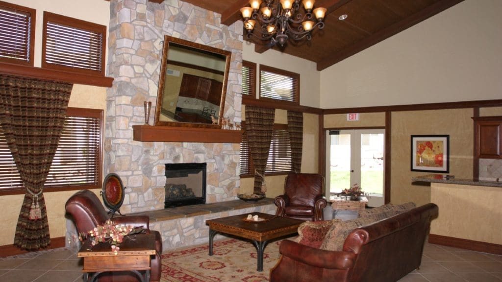 A living room with stone fireplace, two leather chairs, a sofa, a chandelier, rug, and large windows with drapes; kitchen area visible in the background.