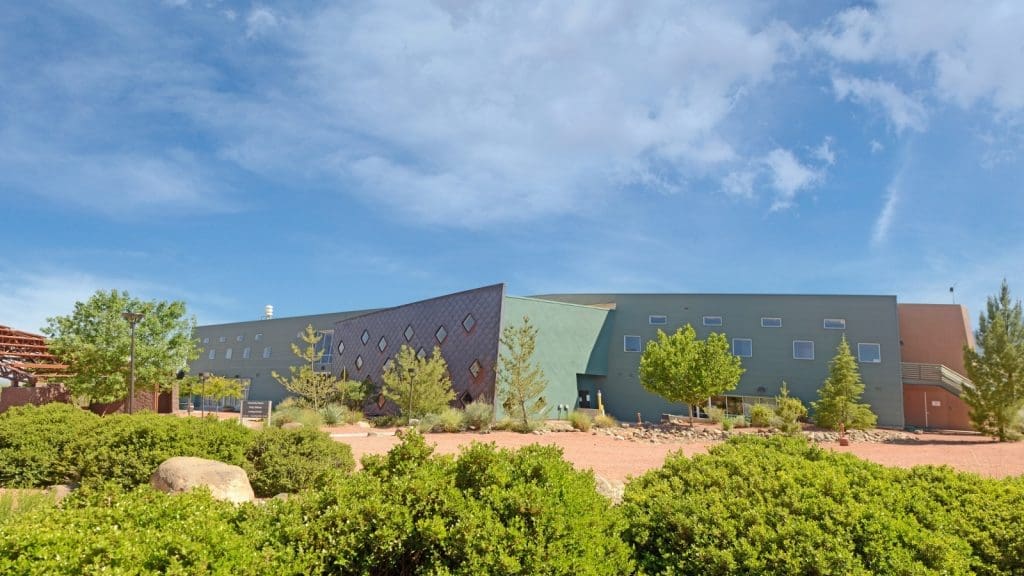 Modern building with geometric design elements, surrounded by desert landscaping with shrubs, rocks, and trees under a partly cloudy sky.