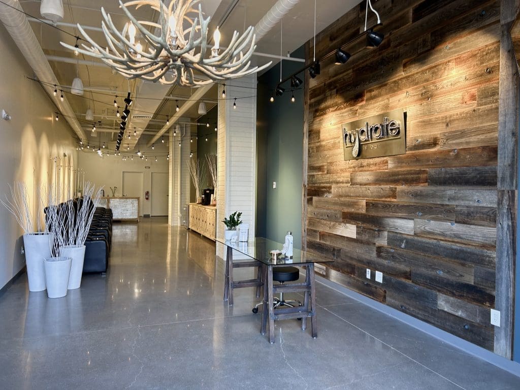 Modern office lobby with polished concrete floors, rustic wood accent wall featuring a "hydrate" sign, antler chandelier, black seating area, and minimalist decor.