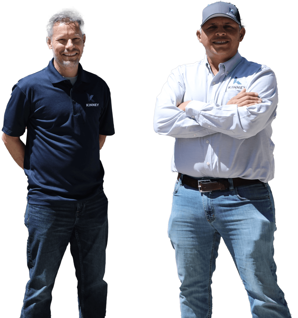 Two men wearing Kinney branded shirts and jeans stand side by side; one has arms crossed, the other has hands behind his back, embodying the casual style that’s all about Kinney.