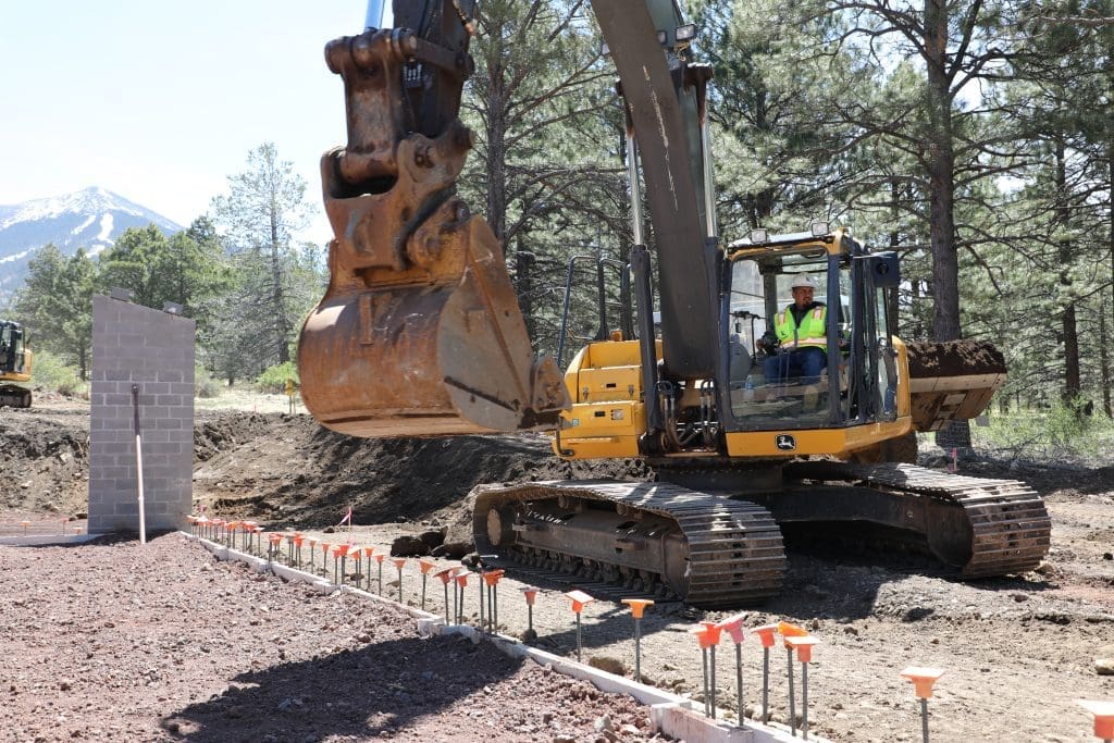 A construction worker operates a large excavator near a partially built concrete wall and rebar at a forested outdoor site.
